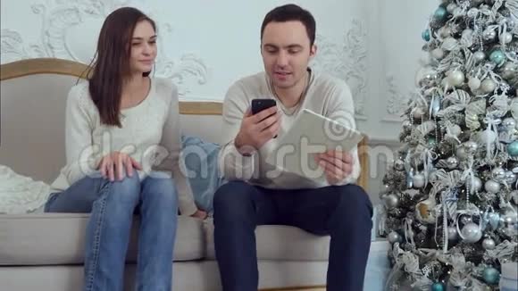 年轻夫妇拿着手机和平板电脑对着镜头说着话在沙发上不寒而栗视频的预览图