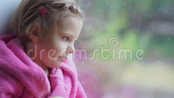 一个穿着粉红色浴袍的小可爱女孩紧紧地盯着窗外坐在窗台上视频的预览图