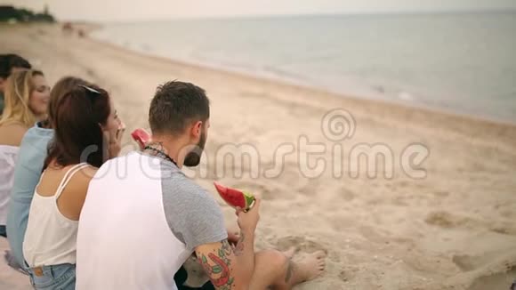 快乐的朋友吃西瓜坐在沙滩度假穿蓝色牛仔裤短裤的男女青年视频的预览图