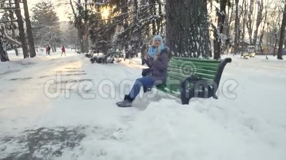 冬天的一天美丽的女孩坐在雪地公园的长凳上视频的预览图
