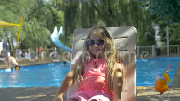 阳光明媚的一天可爱的小女孩戴着太阳镜躺在游泳池背景下的躺椅上视频的预览图