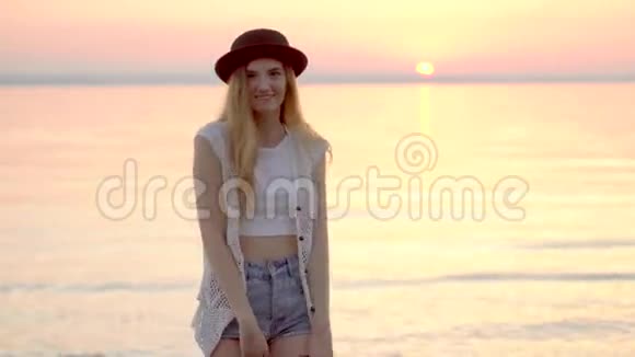年轻的金发女孩戴着帽子在海洋上享受暑假金色的夕阳视频的预览图