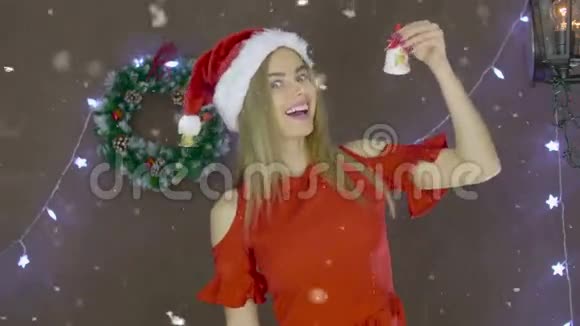 漂亮的金发女孩穿着红裙子戴着圣诞帽在圣诞装饰的背景上敲钟视频的预览图