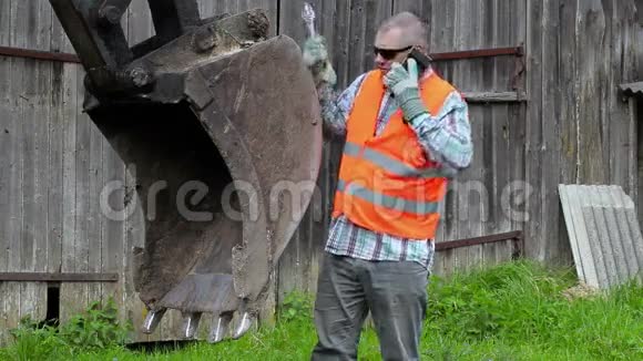 愤怒的工人在拖拉机挖土机铲边用智能电话交谈视频的预览图