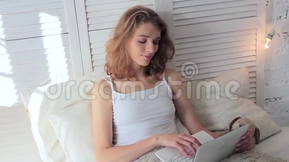 快乐的年轻女人早上躺在床上在笔记本电脑上写着一条信息视频的预览图