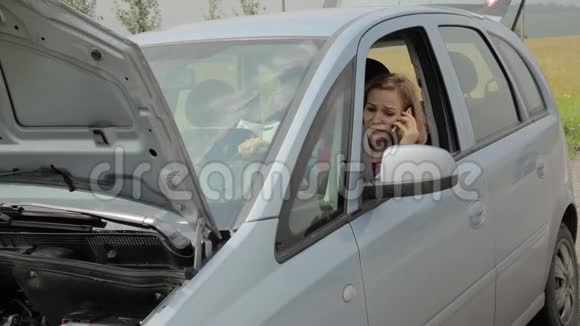 一个金发女孩惊慌失措地坐在她的破车里在乡间的路上开着头罩打着手机视频的预览图