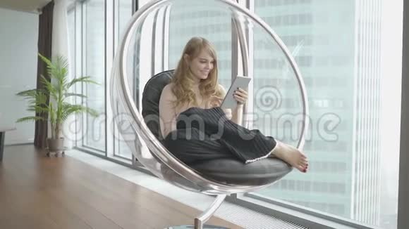 年轻女子在家坐在窗前的现代椅子上用平板电脑工作视频的预览图
