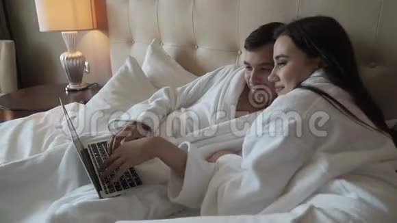 爱一对穿着白大褂的情侣躺在床上休息在MacBook股票视频后面玩得开心视频的预览图