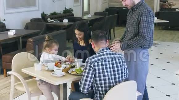 可爱的服务员在智能手机上的咖啡馆里为友好的年轻家庭拍照视频的预览图