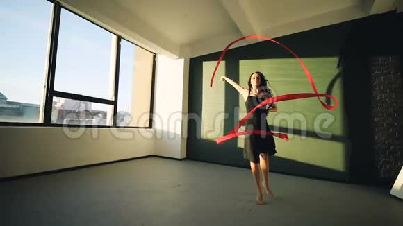 布鲁内特年轻女子舞蹈艺术体操现代用红色丝带穿黑色连衣裙在窗户前太阳视频的预览图