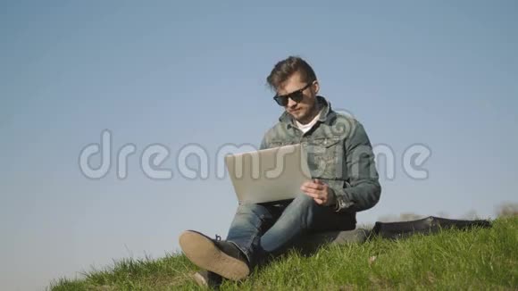 戴着太阳镜的年轻人在公园的草地上坐着笔记本电脑视频的预览图