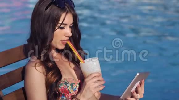 漂亮的女孩用平板电脑和晒太阳附近的游泳池她喝鸡尾酒视频的预览图