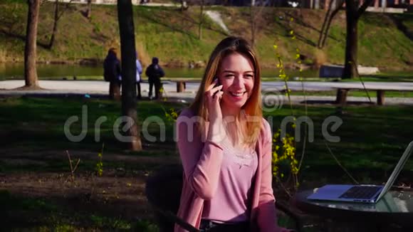 漂亮的女孩坐在公园里拿着笔记本电脑用智能手机说话视频的预览图