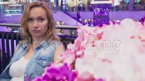一位美丽的年轻女子坐在一个靠近花坛的大型现代购物中心的长凳上视频的预览图