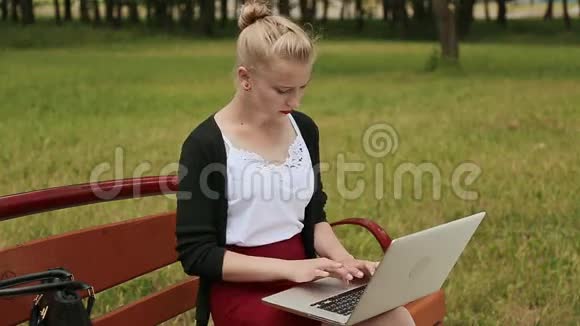 美丽的年轻女孩金发放在公园的长凳上在她的笔记本电脑上工作女孩用笔记本电脑打字侧视视频的预览图
