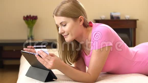 美丽迷人的年轻女子躺在沙发上用信用卡在黑色平板电脑上在线购买视频的预览图