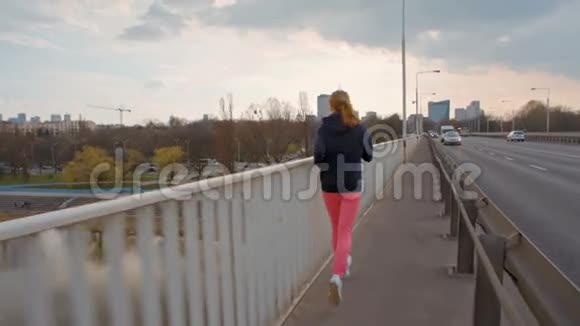 穿着运动服的漂亮年轻女子在城市的一座桥上奔跑的慢动作视频的预览图