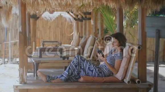 穿着漂亮的女孩躺在巴厘岛热带度假胜地的休息室里年轻女性使用平板电脑和智能手机视频的预览图