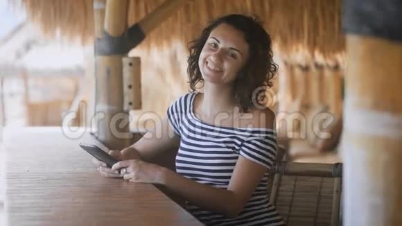 一个晒黑的年轻女孩坐在户外酒吧的草伞下手里拿着一台平板电脑热情地视频的预览图