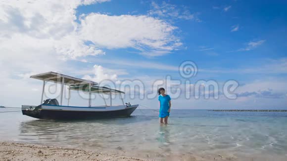 美丽的女孩穿着蓝色的衣服在停泊的船附近的水面上行走苗条的黑发走在沙滩附近视频的预览图