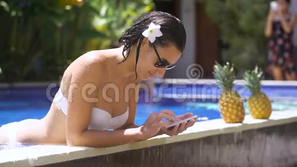 头发上有一朵花的漂亮黑发女人在游泳池边缘使用智能手机穿白衣服洗澡的女孩视频的预览图
