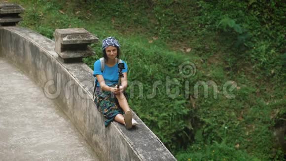 微笑的女旅行者坐在露台上的桥栏杆上手里拿着自拍棒吸引力视频的预览图