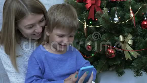 快乐的男孩和他的母亲看着圣诞树附近的智能手机视频的预览图