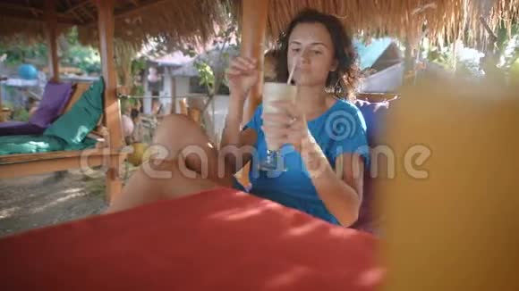 一位黑发的年轻美女穿着一件蓝色的裙子坐在沙滩上坐在稻草下的椅子上视频的预览图
