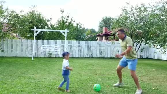 爸爸带着一个四岁的儿子在院子里的绿色草坪上踢球在夏天视频的预览图