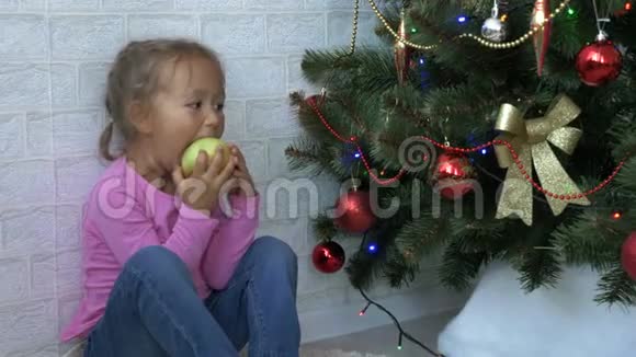 可爱的小女孩坐在地板上在圣诞树旁边吃苹果视频的预览图