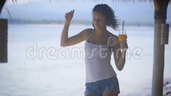 穿着白色背心和牛仔裤短裤的黑发女人正在热带酒吧的海边跳舞享受她的假期视频的预览图