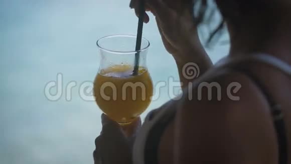 女孩通过吸管喝橙色鸡尾酒并搅拌它人们近距离使用液体透明玻璃视频的预览图