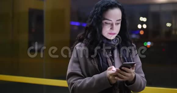 有魅力的女人在夜晚城市的节日灯光的背景下一边使用手机一边穿过街道视频的预览图