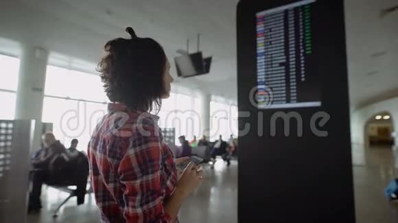 年轻的黑发女人穿着格子衬衫站在机场的信息板附近寻找信息视频的预览图
