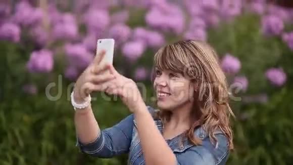 一个年轻的女孩在你的手机上拍照在花丛中自拍视频的预览图