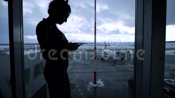 站在机场窗口附近使用智能手机的年轻健康女孩的剪影年轻的商业女性在等待视频的预览图