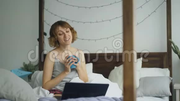 有魅力的开朗女人在家里坐在床上和朋友用笔记本电脑聊天视频的预览图