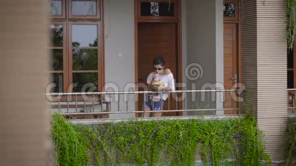 戴太阳镜的女孩来到他房间的阳台上喝椰子带来的异国情调鸡尾酒游客视频的预览图