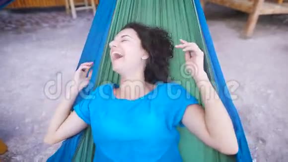 年轻开朗的女孩躺在热带海滩的吊床上掩着脸开心地笑着可爱的蓝色女人视频的预览图