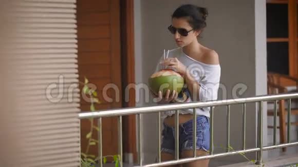 早上戴墨镜的小女孩站在椰子旁边用吸管喝着果汁时尚视频的预览图