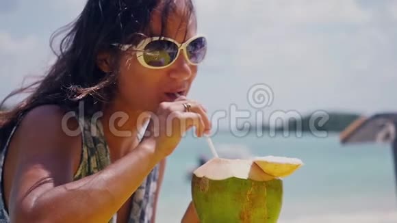 年轻的微笑戴着太阳镜的女人在海滩上享受有趣的假期喝着带吸管的新鲜椰子水慢动作视频的预览图