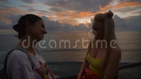 两个快乐的女旅行者在大海的背景下对夕阳谈笑金发和黑发视频的预览图