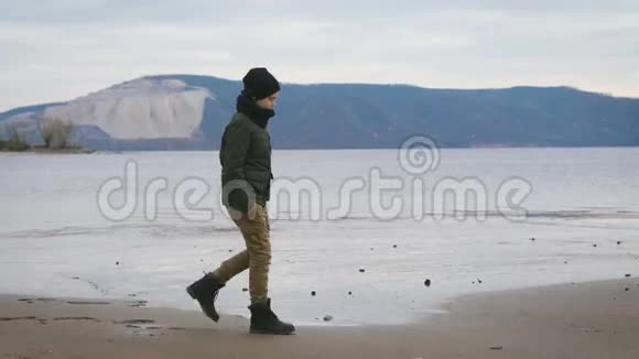 男孩穿着暖和的衣服在海滩上散步它温暖的夹克帽子围巾裤子和靴子浅石色反对视频的预览图