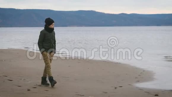 穿着暖和的衣服的男孩正沿着海滨的海岸走着梦里梦里地看着水和山视频的预览图