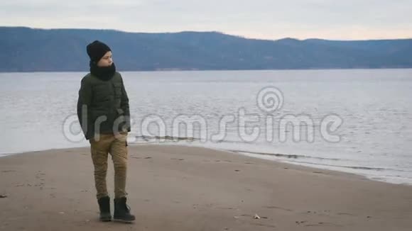 穿夹克戴黑色帽子脖子上戴围巾的男孩在海滩上散步背景下的一种观点美丽的大海视频的预览图