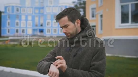 站在橙色和蓝色建筑附近的街道上一个穿着棕色外套的留着胡子的人在检查他的手表丈夫视频的预览图