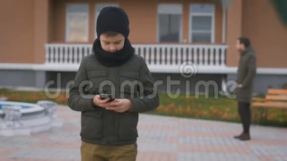 穿着暖色灰色夹克戴着黑色帽子和围巾的男孩正在街上用电话站着在他身后视频的预览图