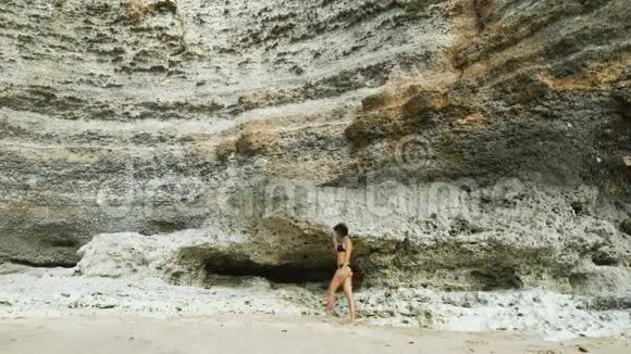 在海滩上穿泳衣的女孩在巨大石壁的背景下她沿着海滩散步她走路视频的预览图