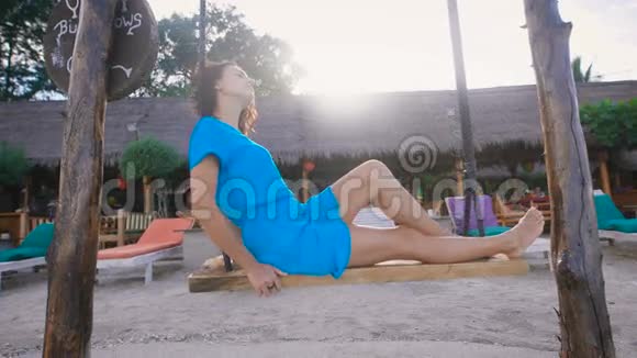 穿着绿松石服装的漂亮的深褐色头发在巴厘岛海滩上荡秋千热带别墅和日光浴床视频的预览图