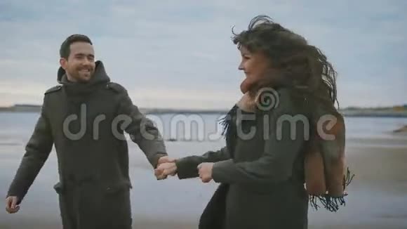 快乐的一对夫妇在沙滩上沿着海滨奔跑手牵着手穿黑色外套和棕色衣服的年轻女子视频的预览图